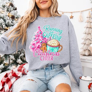 Może przedstawiać: Jasnoszara bluza z kolorową grafiką. Projekt zawiera różową i turkusową choinkę, piernikowego ludzika i kubek kawy z bitą śmietaną. Tekst brzmi "Running on Caffeine and Christmas Cheer."