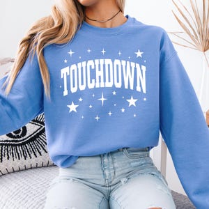 May include: A light blue sweatshirt with the word "TOUCHDOWN" in large white block letters, surrounded by white stars and sparkles. The long-sleeved crew neck top is made of a soft material.