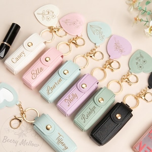 May include: A selection of pastel-coloured lipstick holders, each with a gold keychain and a name engraved on the front. Heart-shaped keychains with floral designs and a black lipstick are also visible.