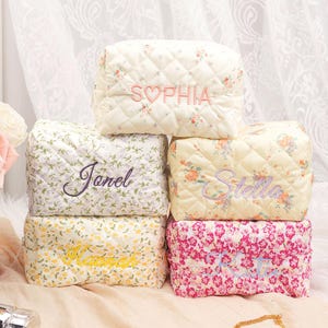 May include: A stack of quilted cosmetic bags with floral patterns in various colors. Each bag is embroidered with a name in a coordinating color. The bags are arranged on a light-colored surface, with a gold necklace and a beauty product visible.