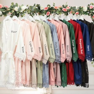 May include: A display of satin robes in a range of colors: white, light pink, blush, champagne, sage green, dusty blue, rose pink, mauve, forest green, burgundy red, navy, and black. Each robe features lace trim and is displayed on a rack.