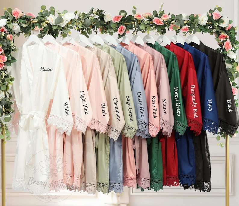May include: A display of satin robes in a range of colors: white, light pink, blush, champagne, sage green, dusty blue, rose pink, mauve, forest green, burgundy red, navy, and black. Each robe features lace trim and is displayed on a rack.