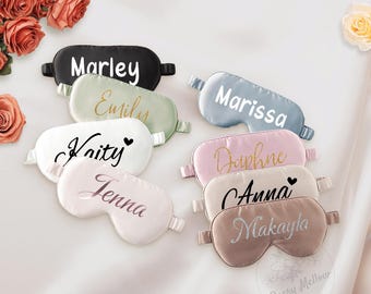 May include: A collection of personalized sleep masks in various colors, including black, sage green, white, light blue, pink, beige, and taupe. Each mask features a name in a contrasting font. The masks are arranged on a white surface with floral accents.