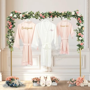 May include: Three satin robes hang on a gold stand adorned with flowers. Two are blush pink with lace trim and the words "Bridesmaid" and "Flower Girl". The third is white with the name "Kayla" embroidered. Slippers with "Kayla Bride" are in front.