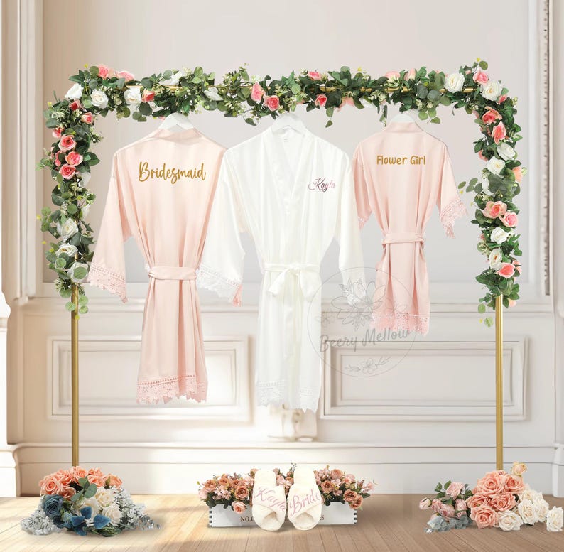 May include: Three satin robes hang on a gold stand adorned with flowers. Two are blush pink with lace trim and the words "Bridesmaid" and "Flower Girl". The third is white with the name "Kayla" embroidered. Slippers with "Kayla Bride" are in front.