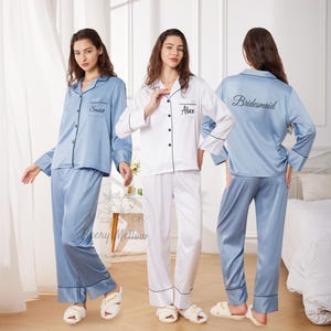 May include: Three sets of satin pajamas in the image. One set is white with black trim, and the other two are light blue with black trim. The light blue pajamas have the names "Scarlett" and "Bridesmaid" embroidered on them. The white pajamas have the name "Alice" embroidered on them.