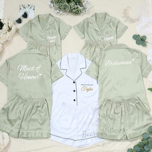 May include: A collection of matching pajama sets in sage green and white. The sets include short-sleeved button-down shirts and shorts. Some shirts are embroidered with titles like "Flower Girl," "Bridesmaid," and "Maid of Honour."