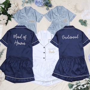 May include: A selection of satin pajama sets in navy, pale blue, and white. The navy set is labelled "Maid of Honour", the pale blue sets have names, and the white set says "Bride". All sets have white piping.