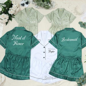 以下が含まれることがあります： 緑と白のサテンのパジャマセットが、花のアクセントとともに展示されています。セットには半袖のボタンダウンシャツとショーツが含まれており、「Maid of Honor」や「Bridesmaid」などの名前やタイトルでパーソナライズされたものもあります。