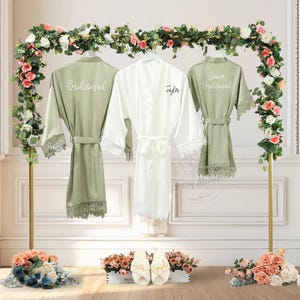 May include: Three satin robes, one white and two sage green, hang on a gold rack decorated with flowers. The white robe is labelled "Taylor", and the green robes say "Bridesmaid" and "Junior Bridesmaid". White slippers with "Bride" and "Taylor" are in front.