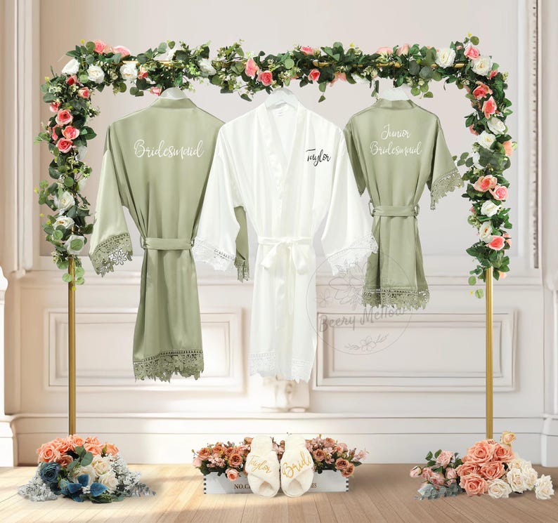 May include: Three satin robes, one white and two sage green, hang on a gold rack decorated with flowers. The white robe is labelled "Taylor", and the green robes say "Bridesmaid" and "Junior Bridesmaid". White slippers with "Bride" and "Taylor" are in front.