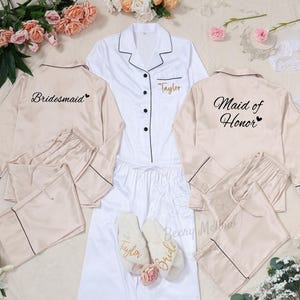 Puede incluir: Una colección de pijamas y accesorios para la fiesta nupcial. Incluye un conjunto de pijama blanco con ribete negro, dos conjuntos de pijama beige y un par de zapatillas blancas. Los pijamas están bordados con "Taylor", "Bridesmaid" y "Maid of Honor".