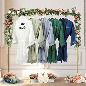 May include: A selection of satin robes in white, sage green, blue, emerald green, and navy blue, each with lace trim and personalised text. The white robe is labelled "Bride" with a crown. The robes hang on a gold rack decorated with flowers.