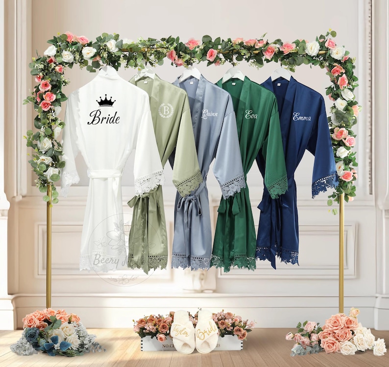May include: A selection of satin robes in white, sage green, blue, emerald green, and navy blue, each with lace trim and personalised text. The white robe is labelled "Bride" with a crown. The robes hang on a gold rack decorated with flowers.