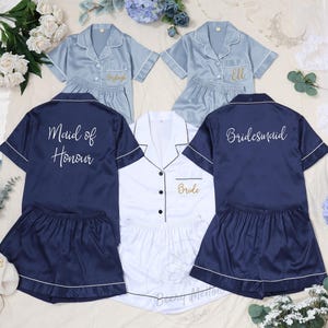 May include: Matching pajama sets in navy blue, light blue, and white, with white piping. The navy set says "Maid of Honour" on the back, and the light blue sets have names embroidered on the front. The white set says "Bride".
