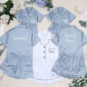 May include: A collection of satin pajama sets in light blue and white. The light blue sets are embroidered with initials and the words "Bridesmaid" and "Maid of Honour". The white set is labeled "Bride". Each set includes a short-sleeved top and shorts.