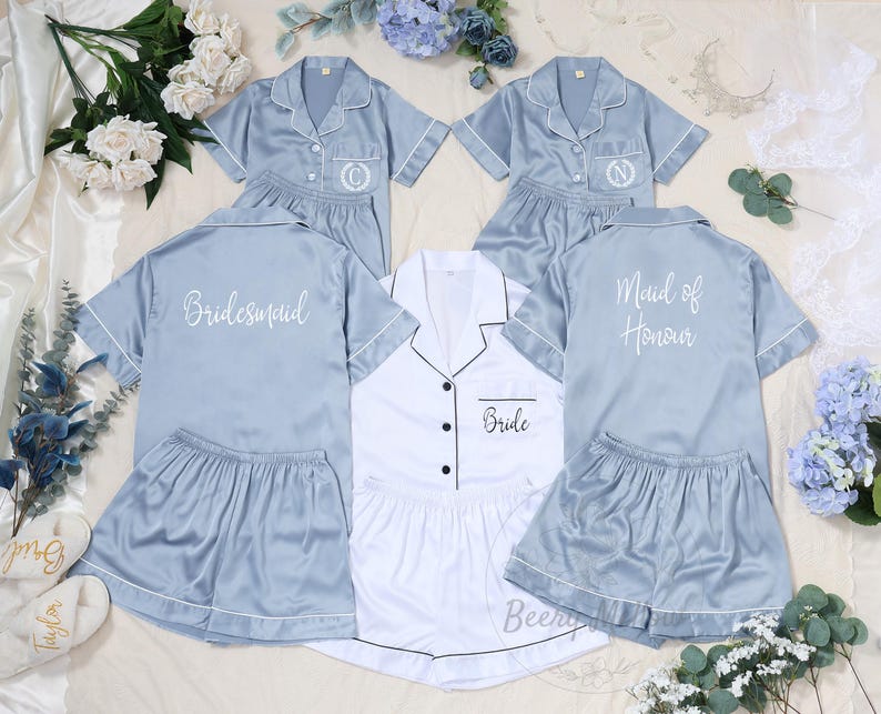 May include: A collection of satin pajama sets in light blue and white. The light blue sets are embroidered with initials and the words "Bridesmaid" and "Maid of Honour". The white set is labeled "Bride". Each set includes a short-sleeved top and shorts.