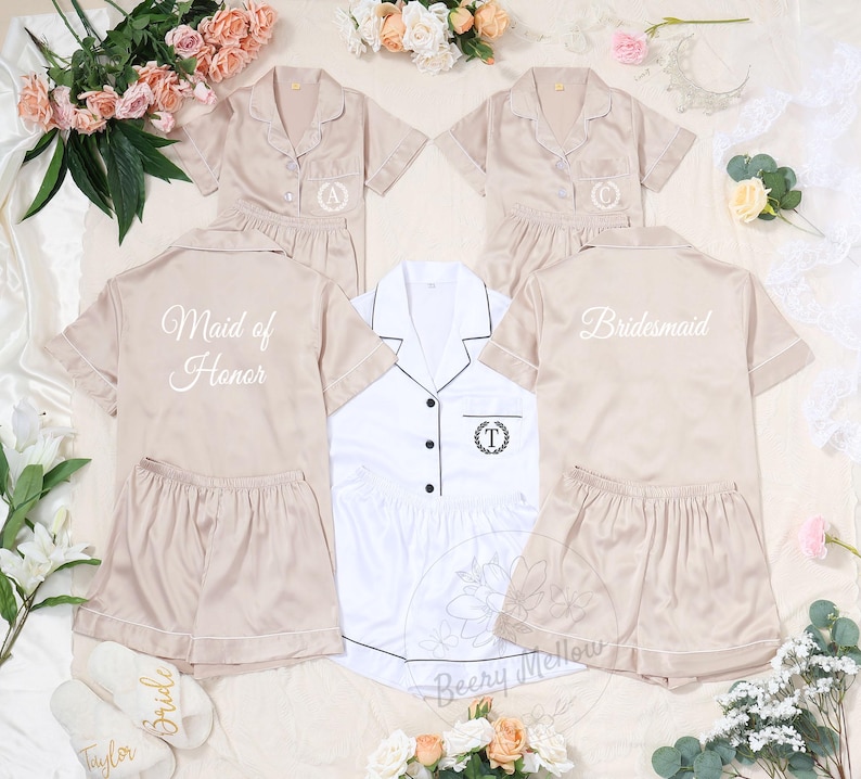 May include: Beige satin pajama sets with white piping, featuring short-sleeved button-up shirts and matching shorts. Some sets are personalized with embroidered initials or titles like "Maid of Honor" and "Bridesmaid".