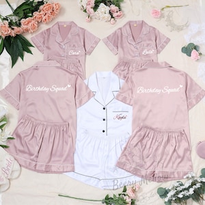 May include: A collection of satin pajama sets in pink and white. The pink sets have "Birthday Squad" printed on the back and personalized names on the front. The white set is also personalized. Matching slippers and eye masks are also included.
