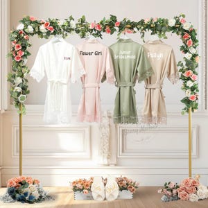 May include: Four satin robes in white, pink, green, and beige hang on a gold rack decorated with flowers. The robes have lace trim and are labeled "Elsie", "Flower Girl", "Junior Bridesmaid", and "Paitlyn". White slippers with "Bride" and "Taylor" are in front.