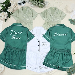 May include: Matching pajama sets in green and white, perfect for a bridal party. The sets include short-sleeved button-down shirts and shorts. Some shirts are embroidered with names and titles like "Maid of Honor" and "Bridesmaid."