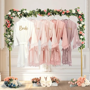 Könnte beinhalten: Präsentation von Brautkleidern in verschiedenen Rosatönen und einem weißen Bademantel, die an einem goldenen Gestell mit Blumen hängen. Der weiße Bademantel trägt den goldenen Schriftzug "Bride". Passende Hausschuhe befinden sich am Boden.