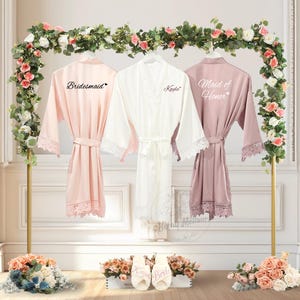May include: Three satin robes in blush, white, and mauve hang on a gold stand, each with lace trim and a matching belt. The robes are labelled "Bridesmaid," "Kayla," and "Maid of Honour." Floral arrangements and slippers with text complete the wedding theme.