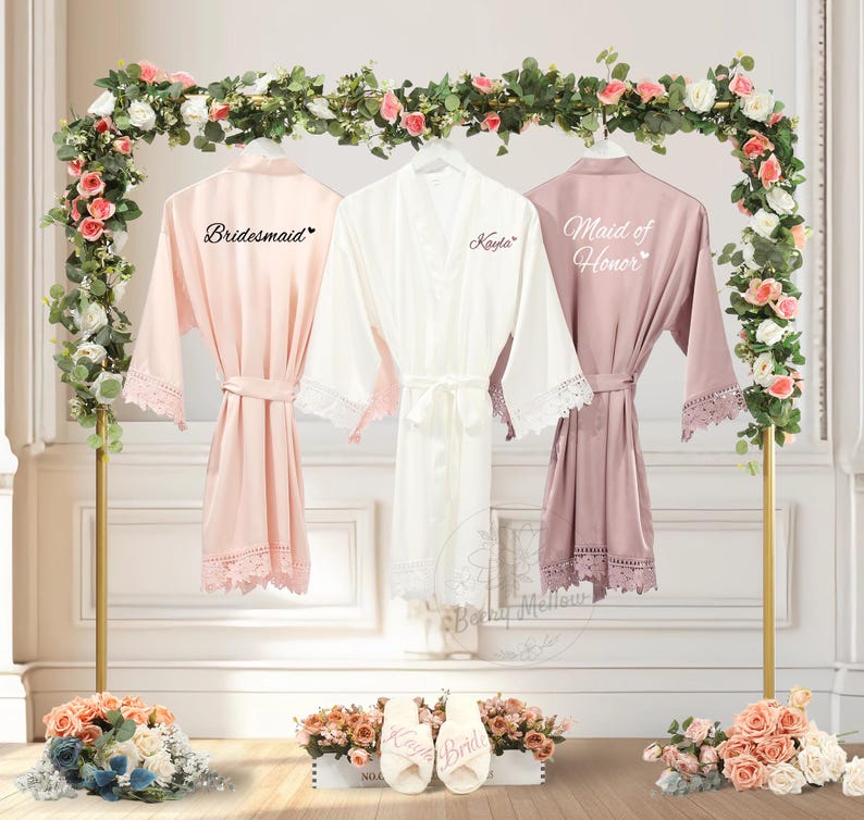 May include: Three satin robes in blush, white, and mauve hang on a gold stand, each with lace trim and a matching belt. The robes are labelled "Bridesmaid," "Kayla," and "Maid of Honour." Floral arrangements and slippers with text complete the wedding theme.