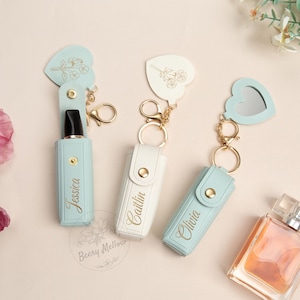 Può includere: Tre porta rossetti personalizzati con portachiavi. I porta rossetti sono in azzurro e bianco, ognuno con un nome in oro. Ogni porta rossetto ha una parte superiore a forma di cuore con un motivo floreale. Uno ha uno specchio.