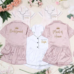 May include: Satin pajama sets in pink and white, perfect for a bridal party. The sets include short-sleeved button-down shirts and matching shorts. Some shirts are personalized with names and titles like "Bridesmaid" and "Maid of Honor".