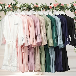 Op de afbeelding: Een verzameling satijnen badjassen in diverse kleuren, waaronder wit, roze, mauve, groen en marineblauw, hangend aan een rek. Elke badjas heeft een bijpassende ceintuur en de kleurnaam is op de mouw gedrukt. De badjassen worden tentoongesteld onder een bloemenboog.