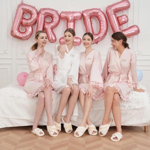 May include: Four people wearing personalised bridal party robes and slippers, sitting on a bed. The robes are pink and white with lace trim. Pink balloons spell out "BRIDE" in the background. The slippers are white with gold text.