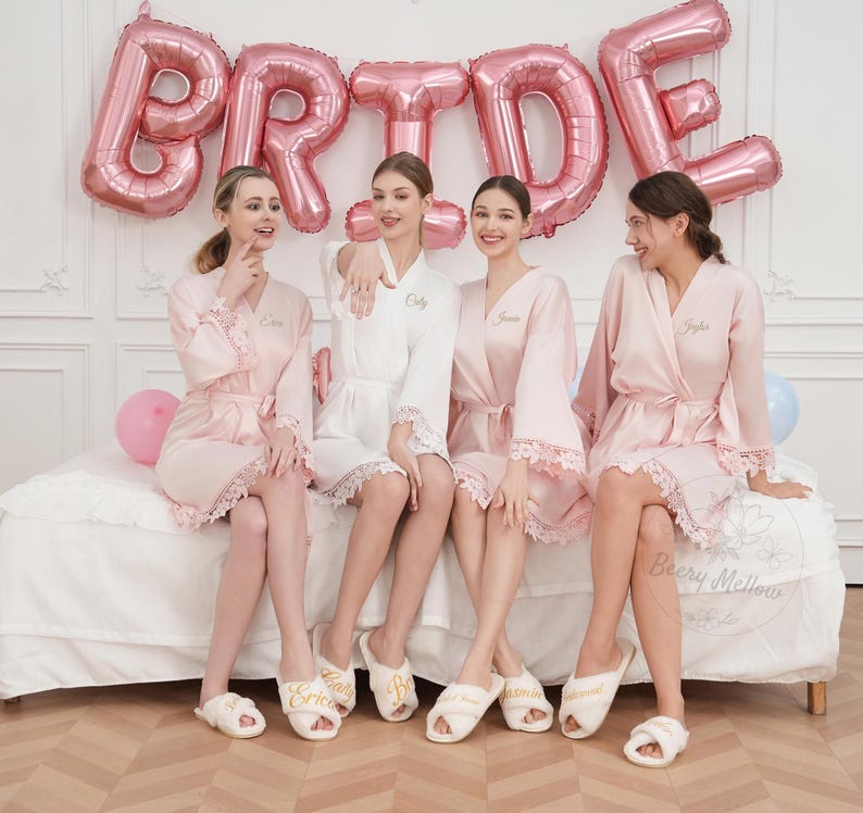 May include: Four people wearing personalised bridal party robes and slippers, sitting on a bed. The robes are pink and white with lace trim. Pink balloons spell out "BRIDE" in the background. The slippers are white with gold text.