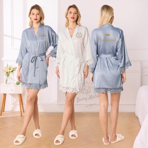 Könnte beinhalten: Drei Frauen tragen Satin-Roben. Eine ist hellblau mit dem Namen "Erika" auf der Vorderseite gestickt. Eine andere ist weiß mit einem Monogramm. Die dritte ist hellblau mit "Bridesmaid" und einer Krone gestickt. Jede Robe hat eine Spitzentrimmung.
