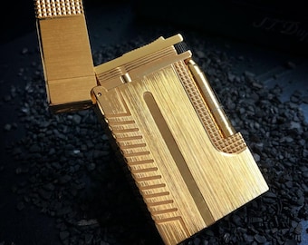 May include: A gold-colored lighter with a textured surface and a rectangular shape. The lighter is open, revealing the interior components. The design includes a grid pattern on the top and a series of parallel lines on the side.