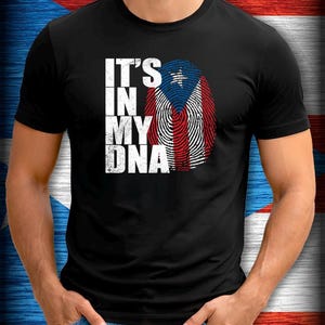 Puede incluir: Camiseta negra con el texto blanco "IT'S IN MY DNA", sobre un diseño de huella dactilar con los colores de la bandera puertorriqueña. La camiseta es de manga corta.