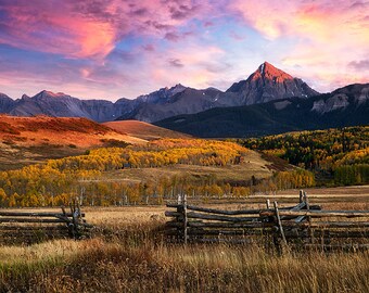 May include: A scenic landscape with a mountain range in the background, a field of golden trees in the foreground, and a wooden fence in the middle ground. The sky is a vibrant pink and orange.