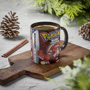Peut inclure: Une tasse à café noire avec l'illustration du jeu "Pokémon Version Rubis". La tasse a une anse noire et est remplie de café chaud, de la vapeur s'en dégage. La tasse est posée sur une planche à découper en bois, avec des bâtons de cannelle et des pommes de pin.