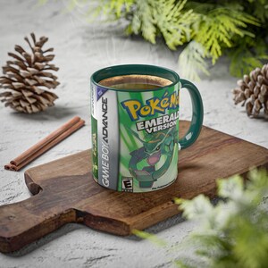 Peut inclure: Une tasse en céramique verte et blanche avec l'illustration du jeu "Pokémon Version Émeraude". La tasse a une anse et un bord verts et est remplie d'un liquide foncé. La tasse est posée sur une planche à découper en bois.