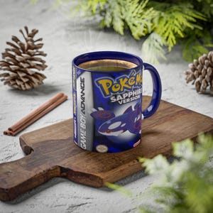 May include: A blue ceramic mug with the "Pokémon Sapphire Version" game cover design. The mug has a blue handle and contains a hot drink. It sits on a wooden board, with cinnamon sticks and pine cones nearby.