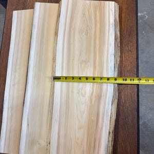 May include: Three unfinished wooden planks with natural edges, light brown in color, are displayed. A yellow measuring tape shows a length of approximately 12 inches. The wood grain is visible, and the planks are on a dark wooden surface.