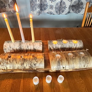 May include: Birch log candle holders with lit candles. The candle holders are made from natural birch logs and hold tall, slender candles and small tea lights. The candles are lit, casting a warm glow. The arrangement is on a wooden table.