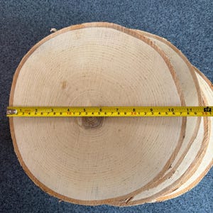 May include: A stack of four natural wood slices, each with a diameter of approximately 10 inches. The wood is light brown and has a natural grain pattern. A yellow measuring tape is placed on top of the wood slices.