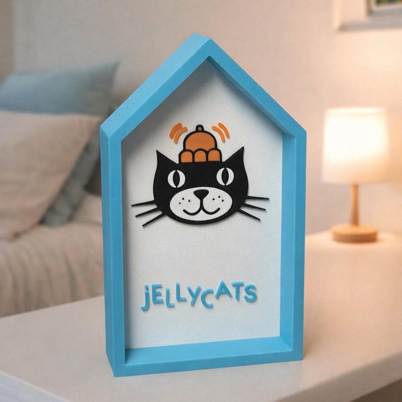 May include: A blue house-shaped frame with a black cat illustration wearing an orange hat. The cat has white eyes and a smiling mouth. The word "JELLYCATS" is written in blue letters at the bottom of the frame.