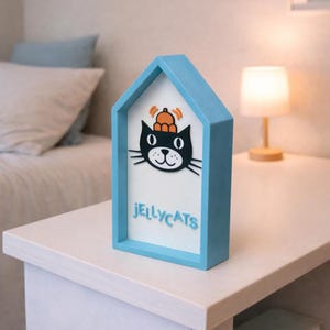 May include: A blue house-shaped decorative item with a black cat graphic wearing a hat. The word "JELLYCATS" is written in blue below the cat. The item is on a white table.
