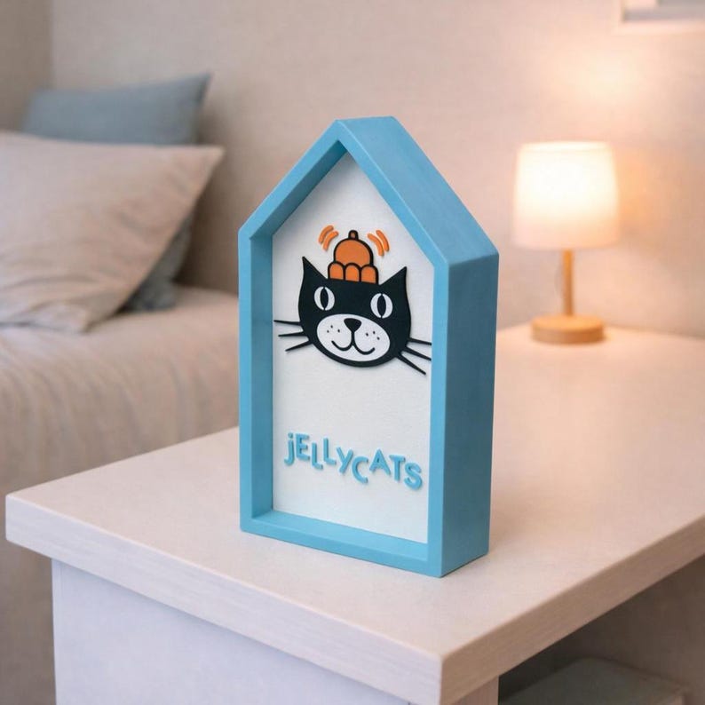 May include: A blue house-shaped decorative item with a black cat graphic wearing a hat. The word "JELLYCATS" is written in blue below the cat. The item is on a white table.