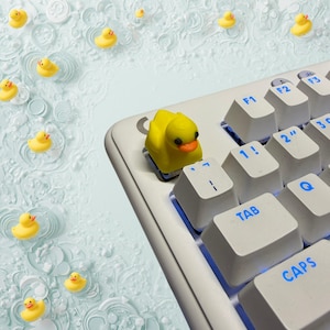 May include: A yellow rubber duck keycap sits on a white keyboard, surrounded by other rubber duckies. The keycap has an orange beak and black eyes. The keyboard keys are labelled with blue text.