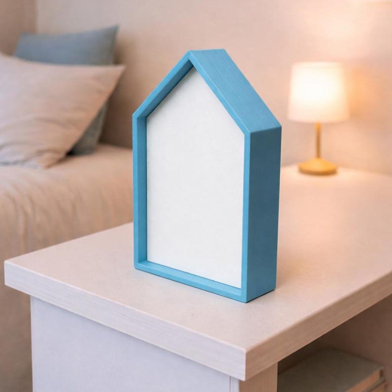 May include: A blue house-shaped picture frame with a white interior. The frame is made of wood and sits on a white table. A lamp and bed are in the background.