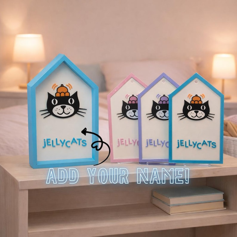 May include: Four house-shaped decorative signs with a black cat graphic wearing a hat, the text "JELLYCATS", and the words "ADD YOUR NAME!". The signs have blue, pink, and purple frames. The signs are on a wooden nightstand.