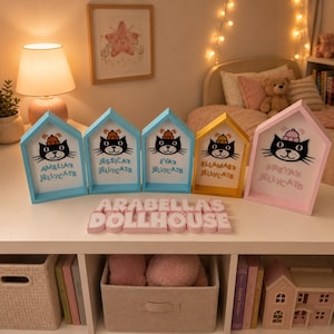 May include: A set of five colorful, house-shaped decorative signs. Each sign features a cartoon cat design with a name below. The names are Amelia's, Jessica's, Eva's, Ellamae's, and Freya's. The text "ARABELLAS DOLLHOUSE" is below the signs.